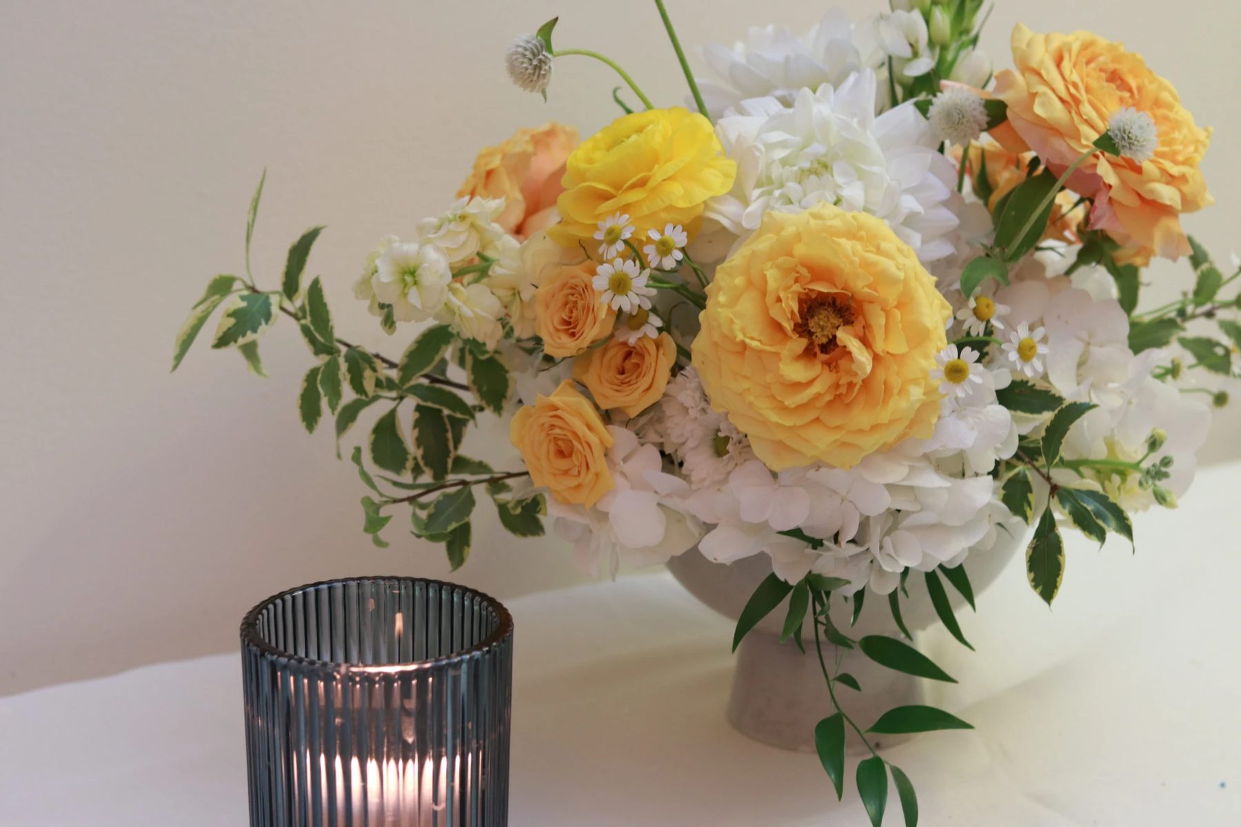 Golden garden rose, yellow ranunculus, white dahlia and hydrangea arrangement in a pale footed bowl beside a votive candle