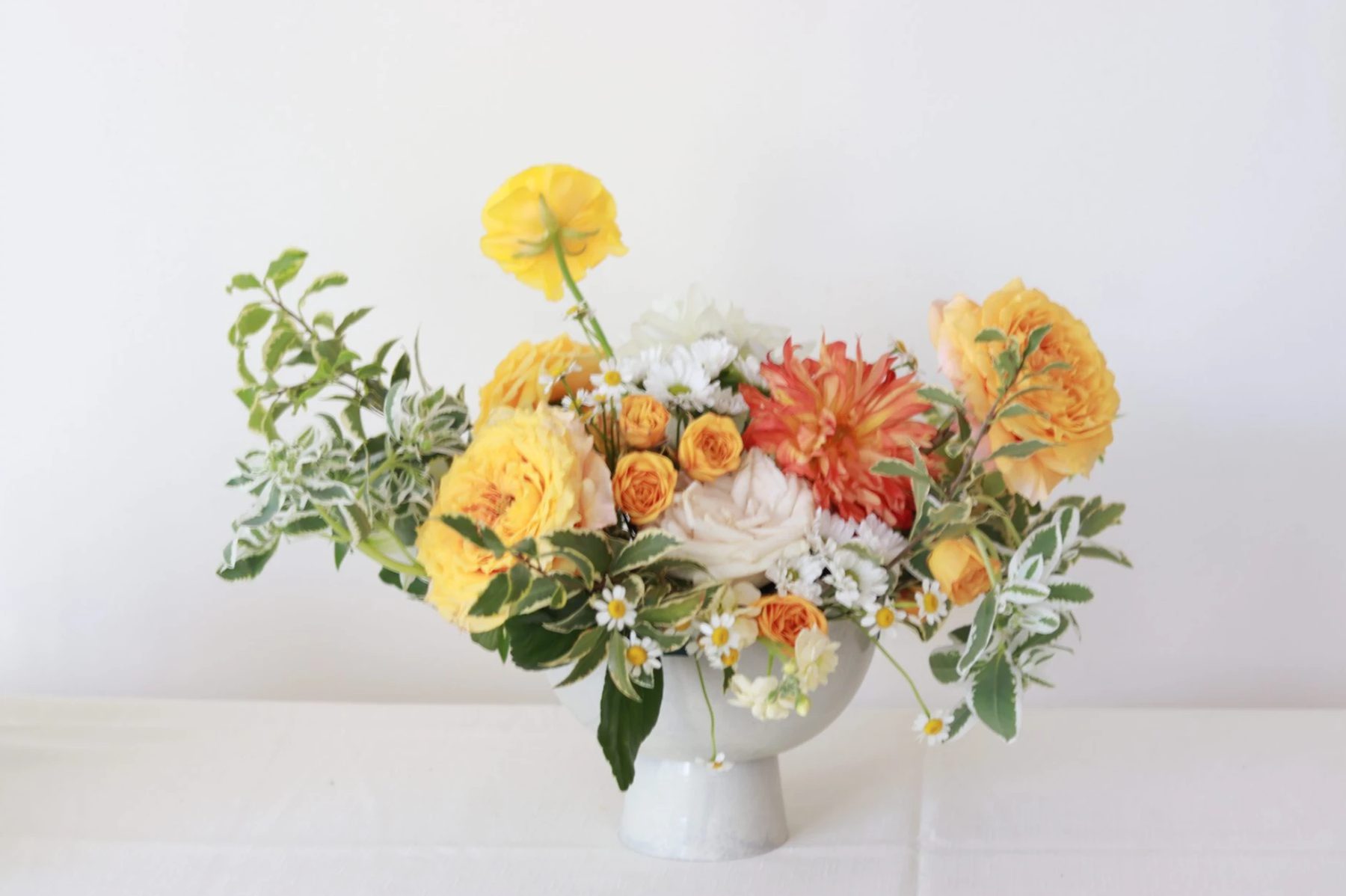 Yellow garden rose, orange dahlia, and daisy arrangement in a pale footed bowl