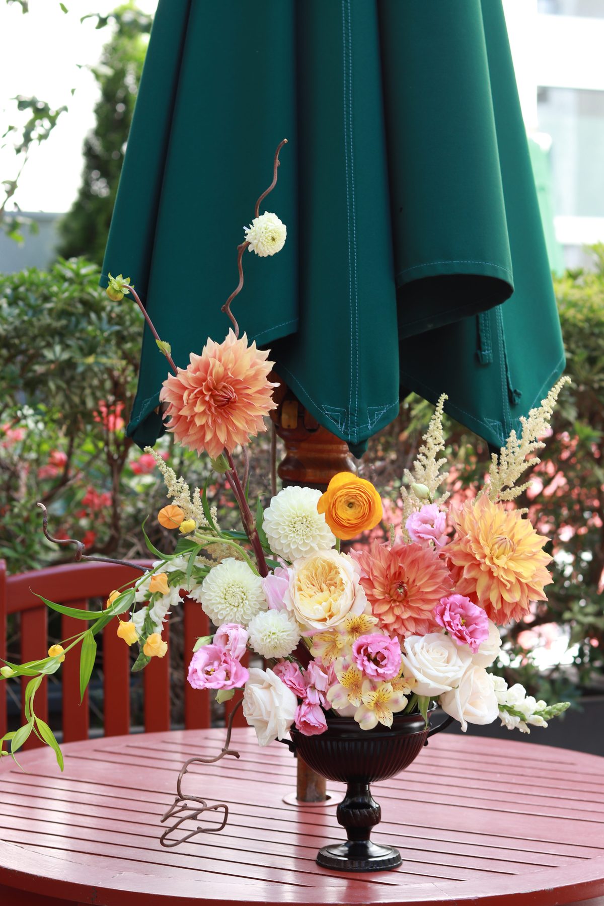 Colorful dahlia, rose, and ranunculus arrangement in a dark urn on a patio table under a green umbrella