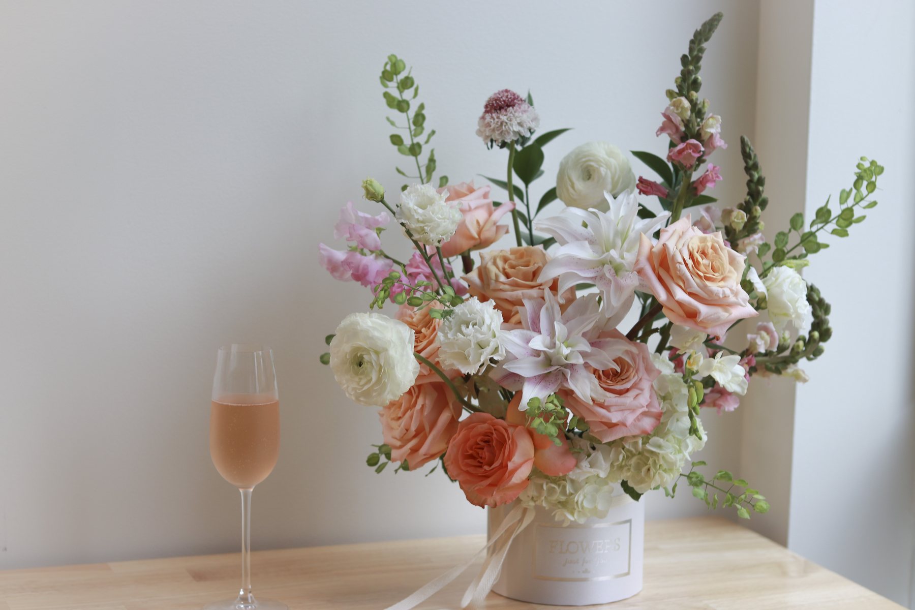 Peach rose and ranunculus arrangement in a white flower box, styled beside a glass of rosé by a window