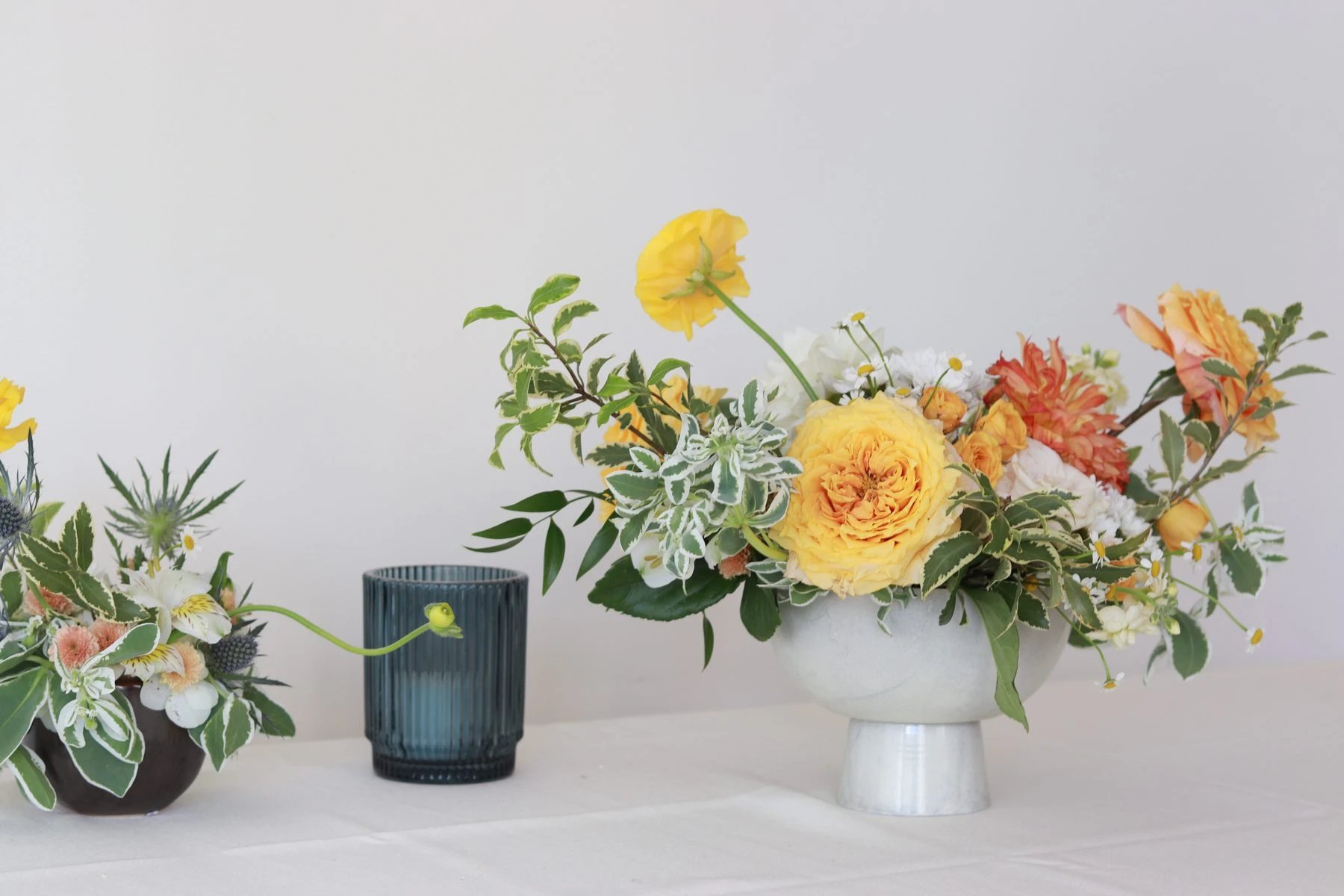 Yellow garden rose, orange dahlia, and ranunculus arrangement in a pale footed bowl beside a blue glass votive