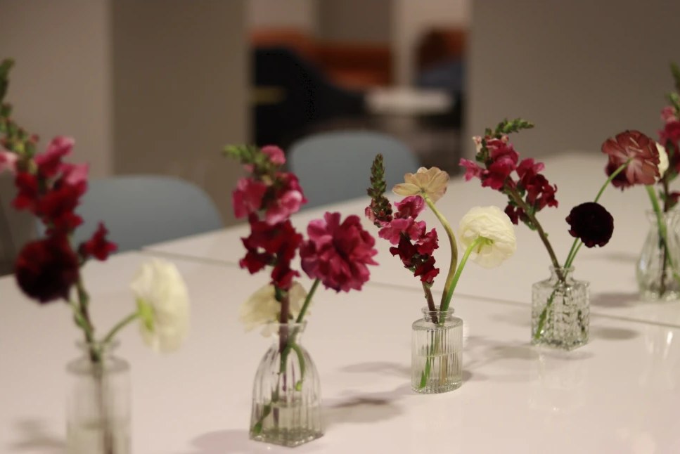 Row of small glass bud vases with burgundy snapdragon, carnation, and white ranunculus on a white table