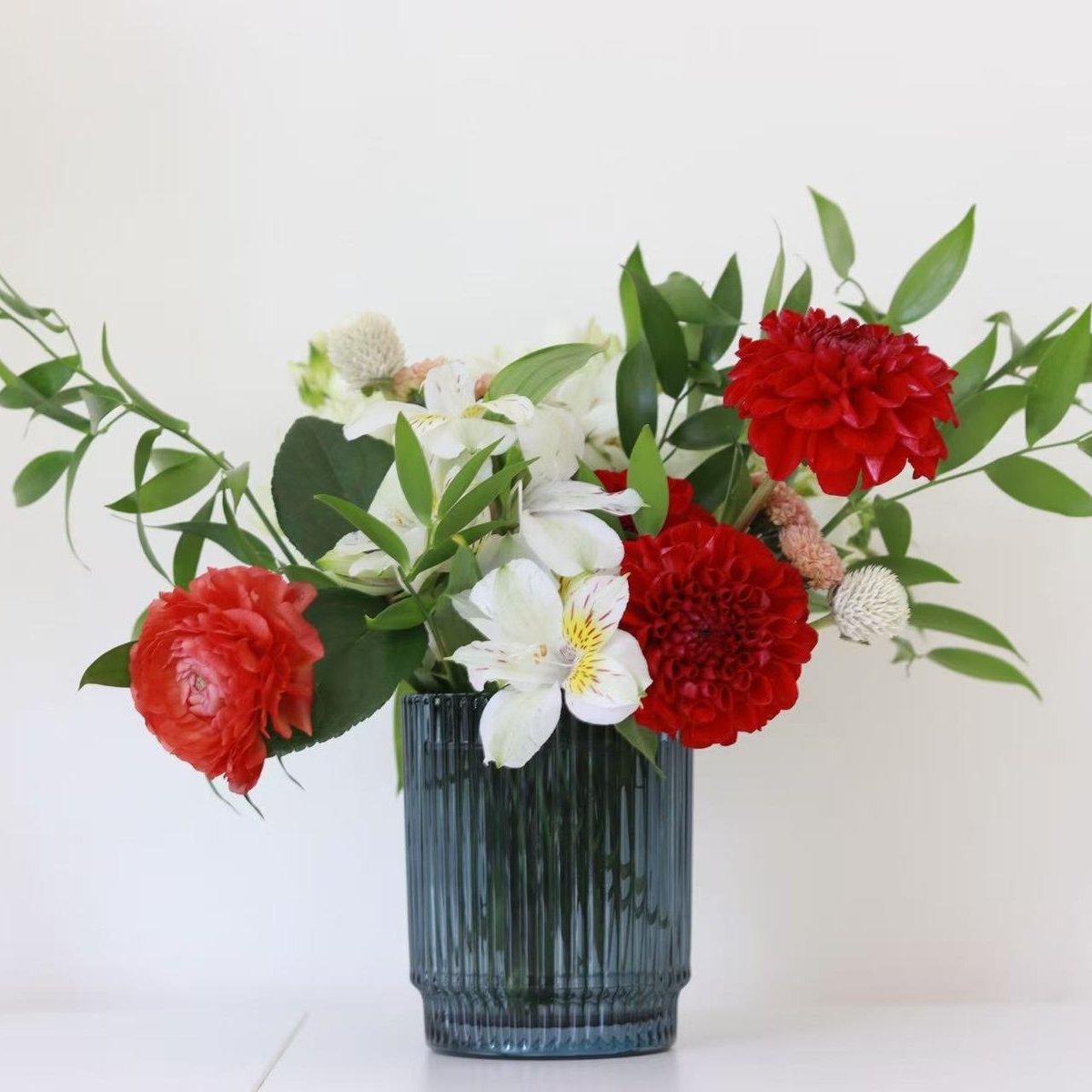 Red dahlia, ranunculus, and white alstroemeria with greenery in a ribbed blue-grey vase