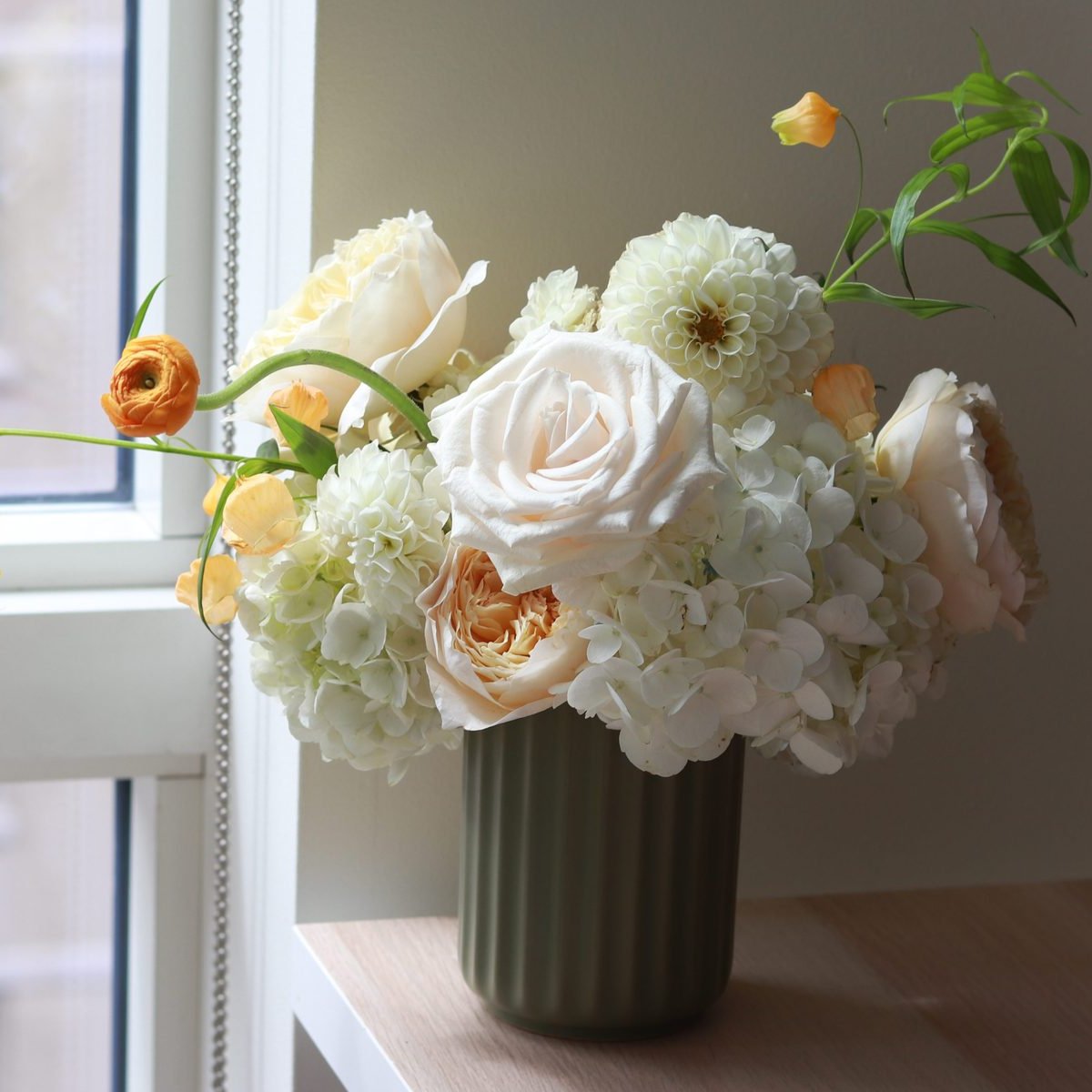 Ivory rose, white hydrangea, and orange ranunculus in a sage green ribbed vase by a window