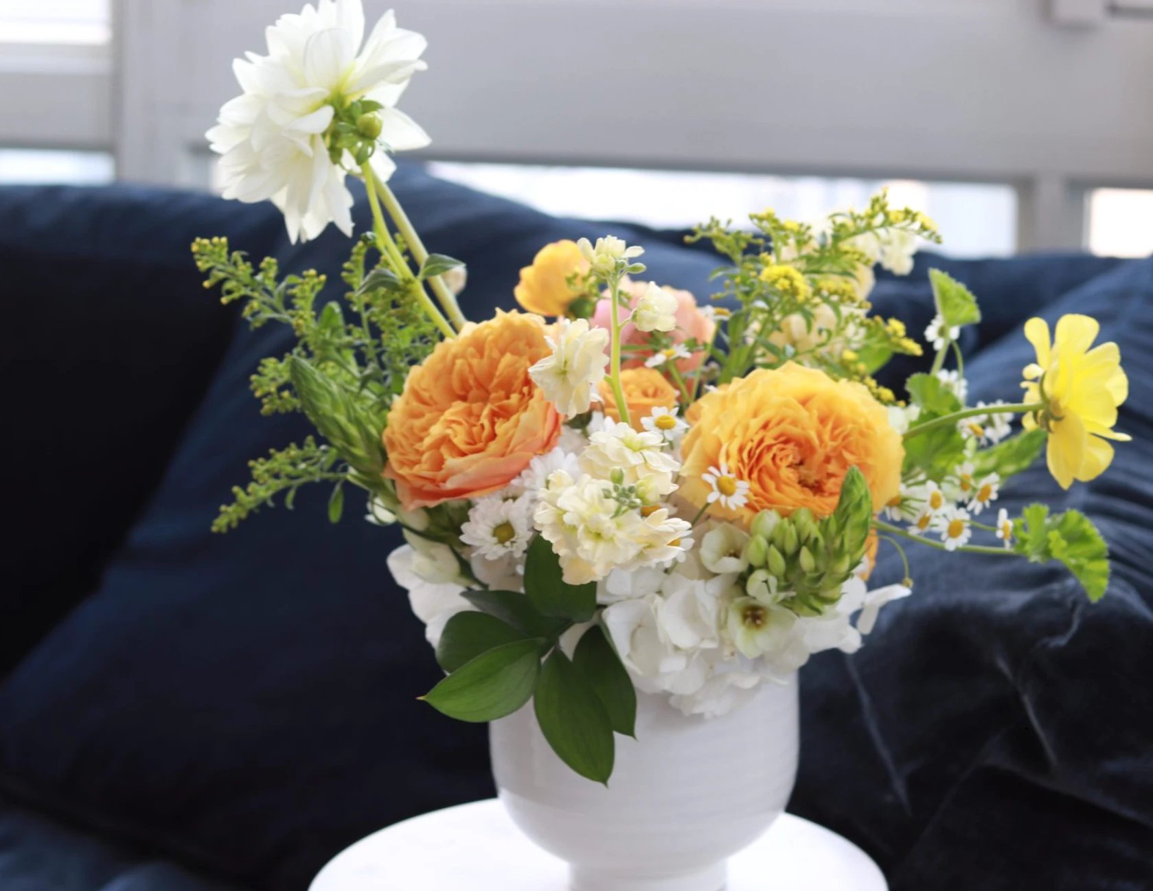 Orange and yellow garden roses, white dahlia, and hydrangea in a white footed vase on a navy sofa