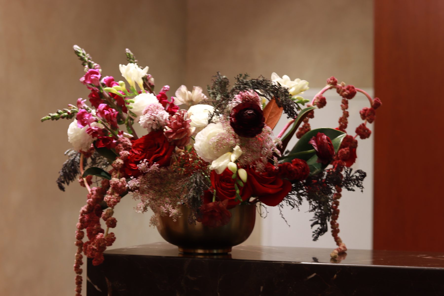 Burgundy rose, ranunculus, and amaranthus arrangement in a brass bowl on a marble lobby console